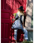 Person carrying navy bandana print tote bag leaning against a red door holding a green cup, wearing a black hat, scarf, and blue jeans.
