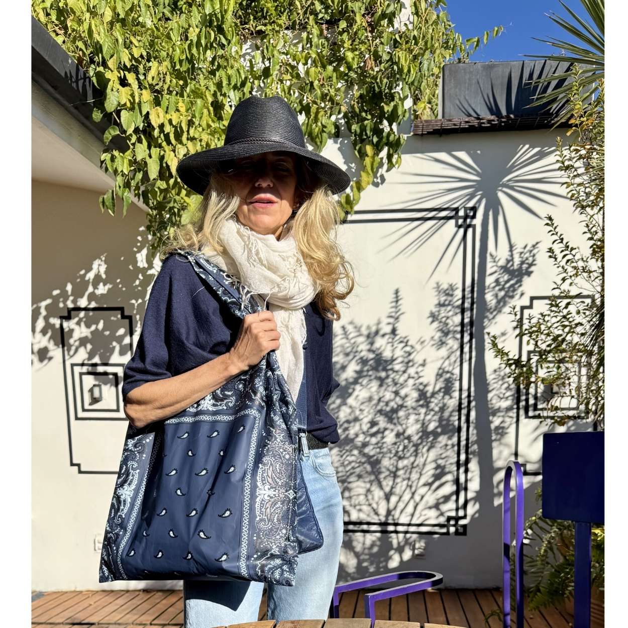 Woman holding a navy blue bandana print tote bag wearing a black hat and scarf, standing outdoors with a building and trees in the background.