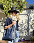 Woman holding a navy blue bandana print tote bag wearing a black hat and scarf, standing outdoors with a building and trees in the background.