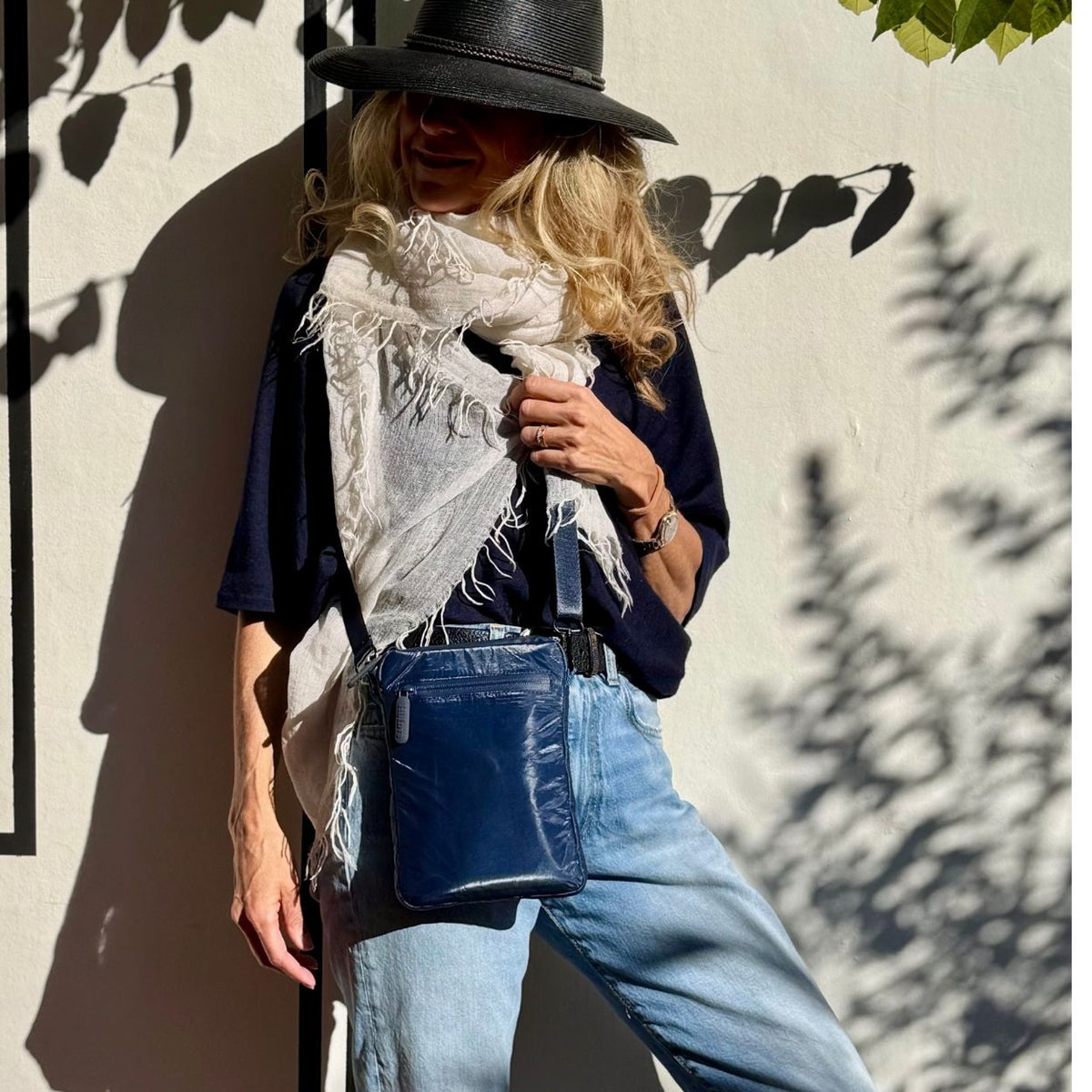 Woman wearing a navy blue crossbody purse leaning against a cement wall with shadows of plants reflected on the wall.