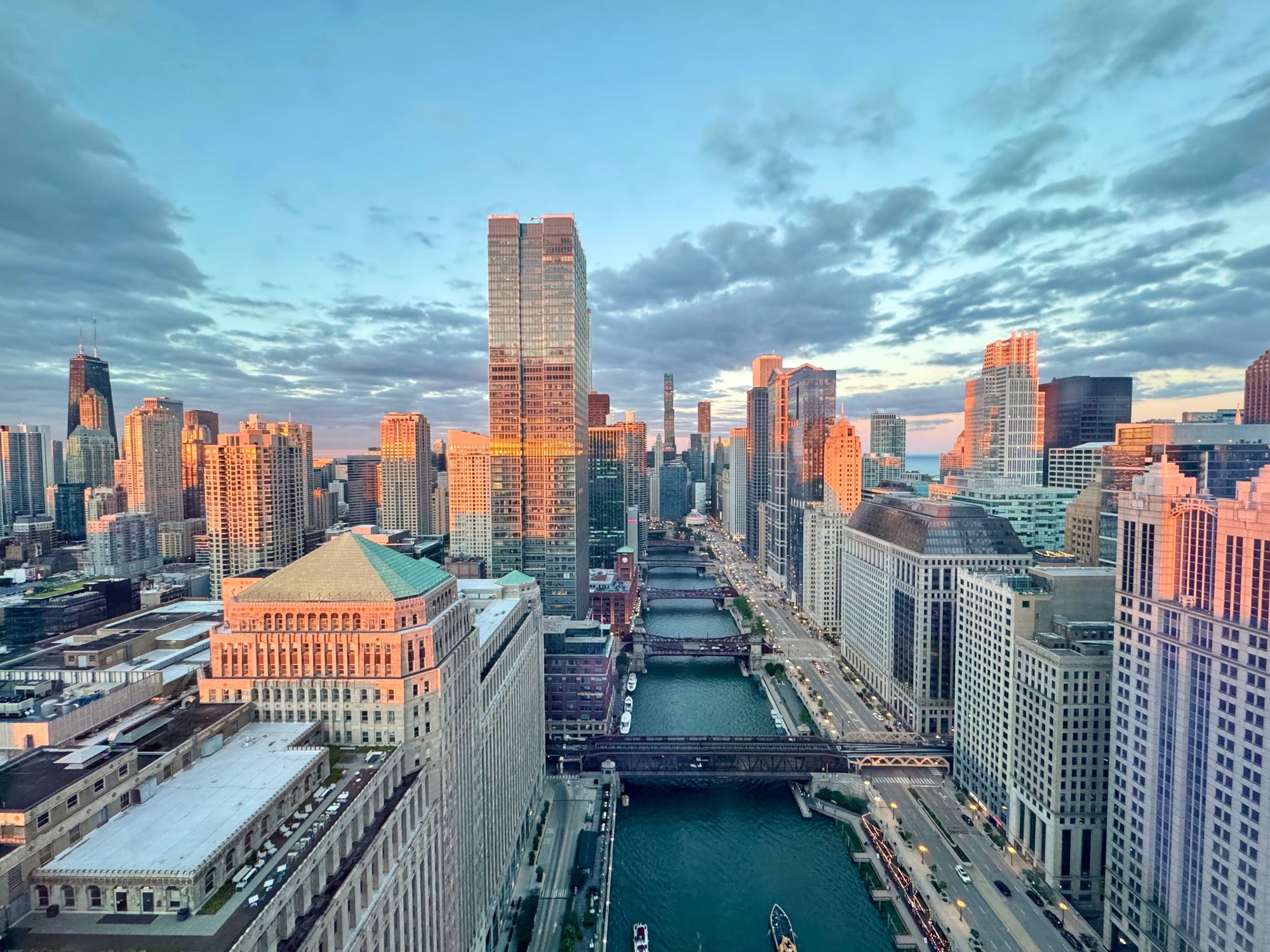 Aerial view of the Chicago skyline with the Chicago River running through the center, surrounded by tall glass skyscrapers and historic buildings under a blue sky with warm sunset light.