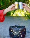 Two people holding drinks above a butterfly-patterned cooler bag with a blurred green background.