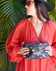 Woman in a red dress holding a black clutch with butterfly designs against a green leafy background