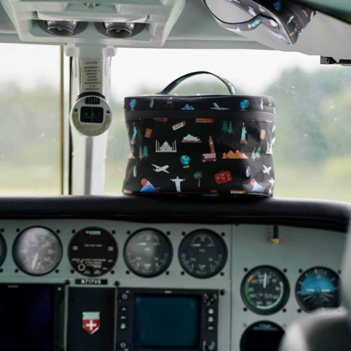 A black travel-themed lunch case with a colorful cartoon print sits on the dashboard of an airplane cockpit.
