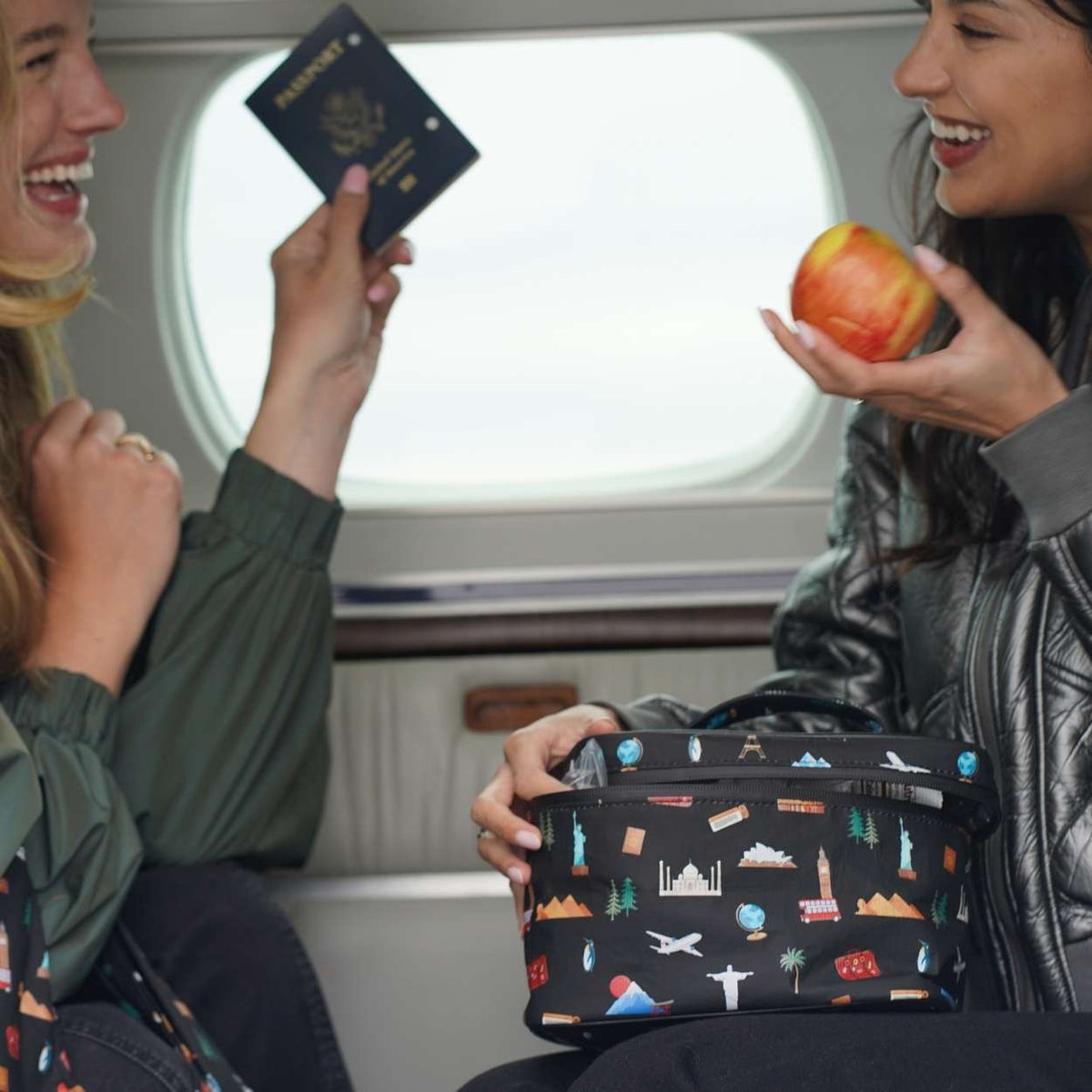 Two women smiling on a plane, holding a black travel-themed lunch bag with a colorful world landmark print. One woman holds a passport, and the other holds an apple.