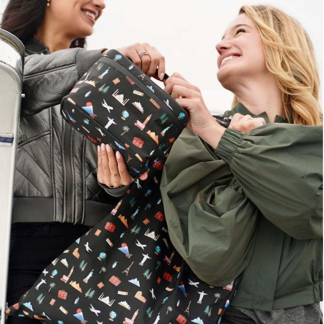 Two women smiling and holding a black reusable tote bag and lunch box with a colorful travel-themed pattern, set against a bright, overexposed outdoor background.
