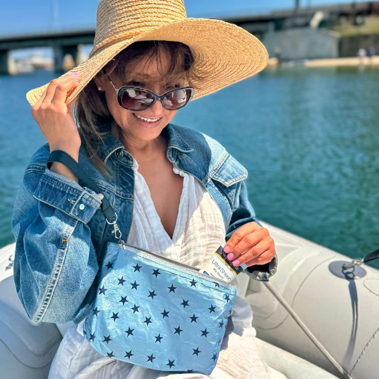 Woman on boat grabbing sunscreen from cosmetic zipper pouch light blue shimmer myriad stars with detachable navy wristlet strap