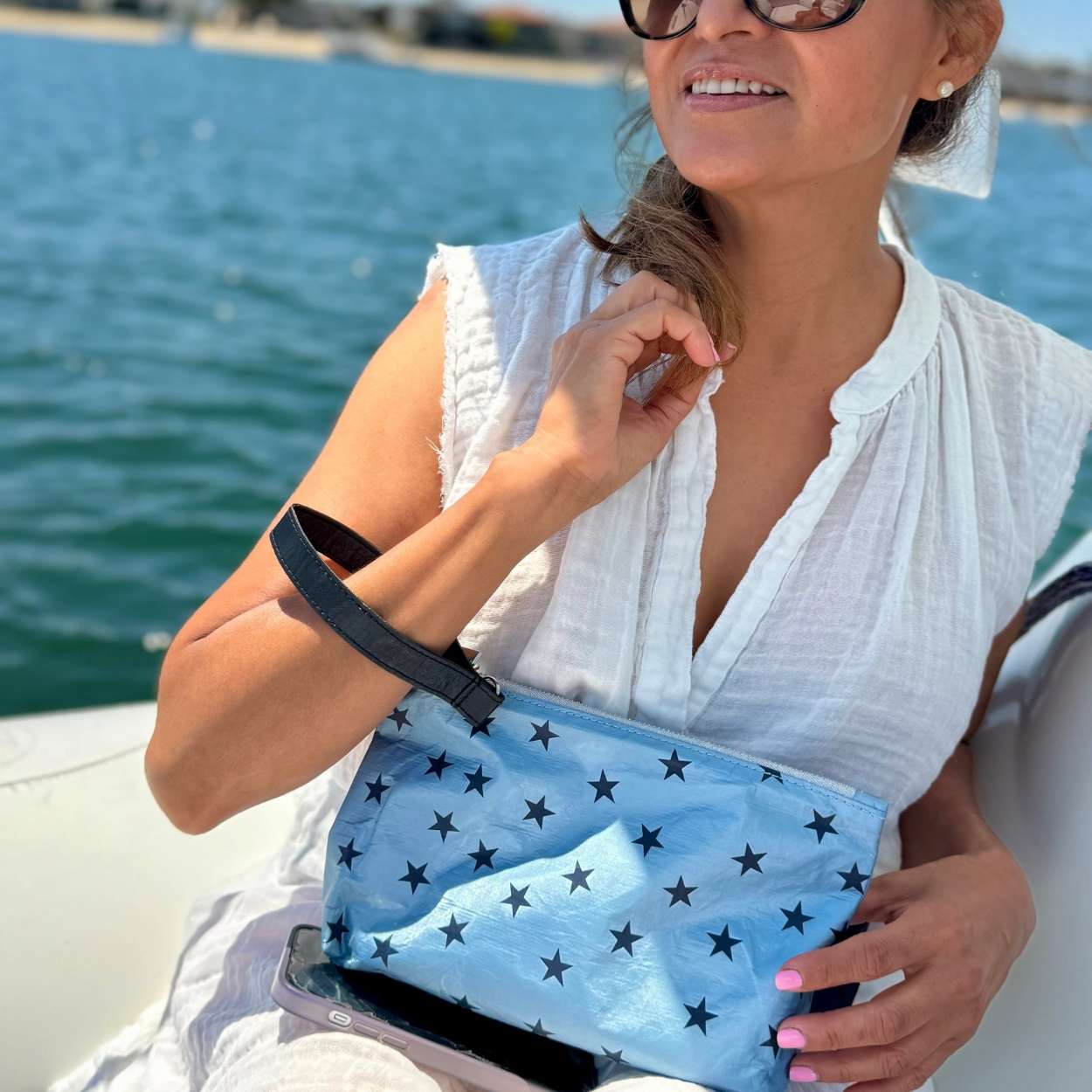 Woman on boat holding cosmetic zipper pouch light blue shimmer myriad stars with detachable navy wristlet strap