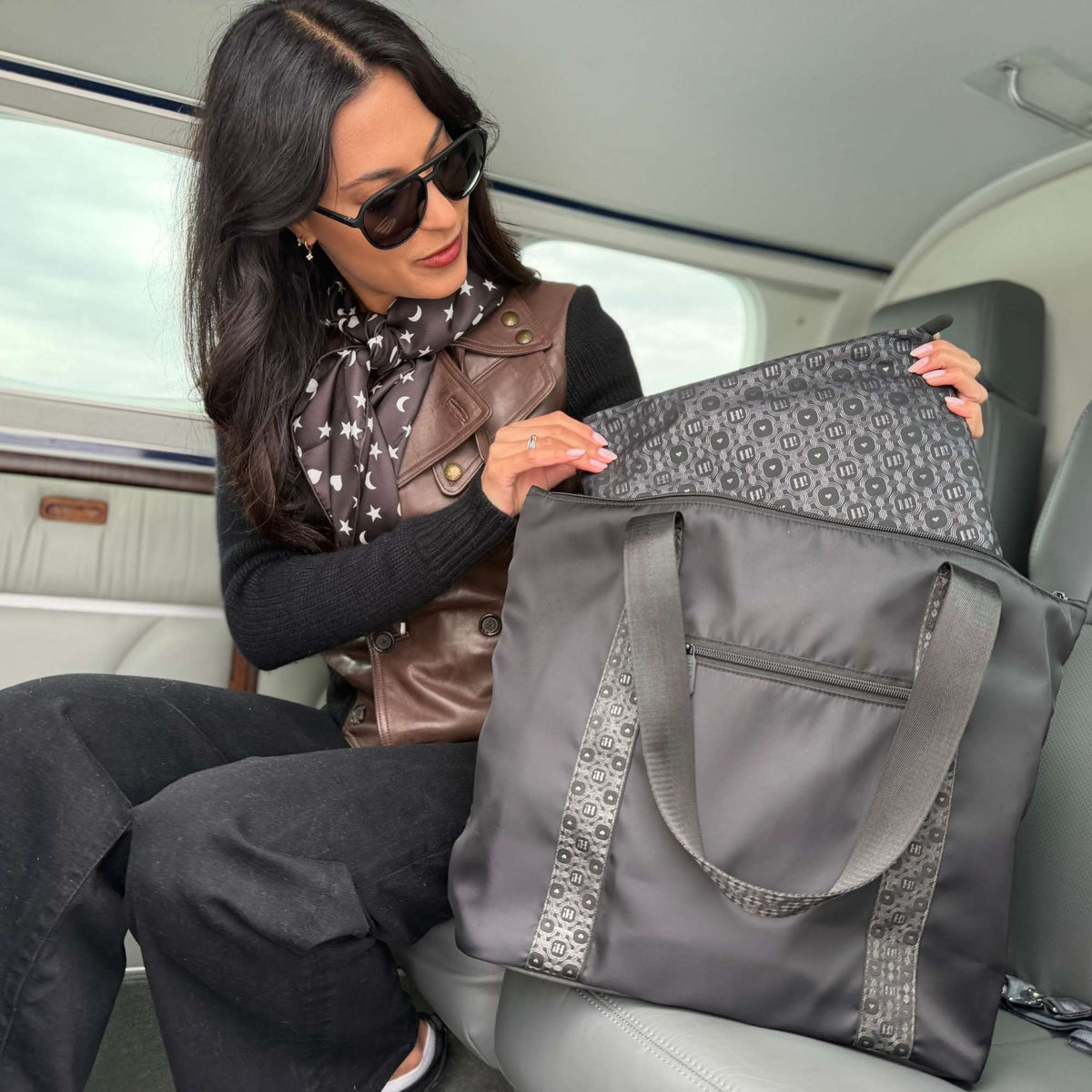 A woman wearing sunglasses and a scarf sits inside a plane, holding open a large black signature tote with patterned fabric lining and handles with a matching medium pouch
