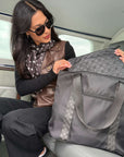 A woman wearing sunglasses and a scarf sits inside a plane, holding open a large black signature tote with patterned fabric lining and handles with a matching medium pouch