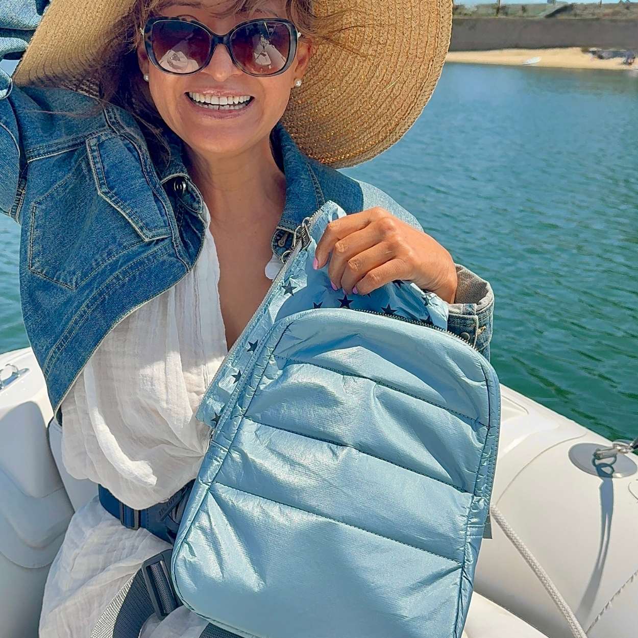 Woman on boat holding crossbody backpack in shimmer light blue and cosmetic zipper pouch in shimmer light blue with myriad of stars