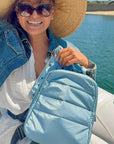 Woman on boat holding crossbody backpack in shimmer light blue and cosmetic zipper pouch in shimmer light blue with myriad of stars