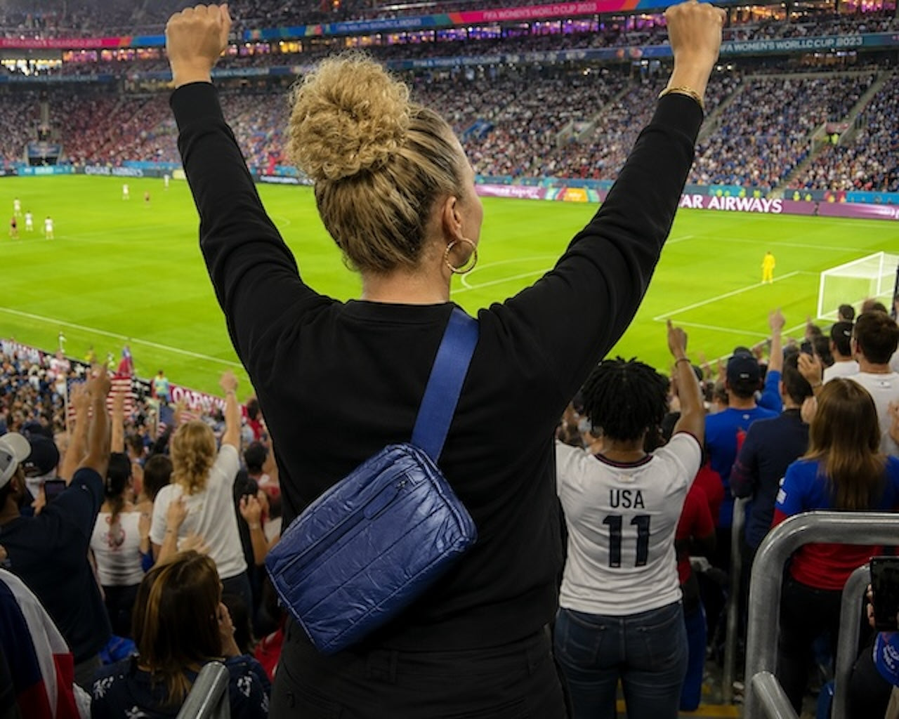Women in the stands of a soccer game cheering and wearing a navy crossbody belt bag.