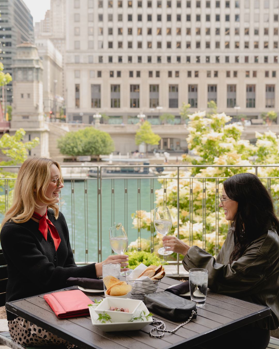 Two women sitting at a wooden outdoor table on a balcony in Chicago, clinking wine glasses. The setting is in a city with a river and large buildings in the background, with flowers and greenery in the foreground. A red and black chain purse on the table.
