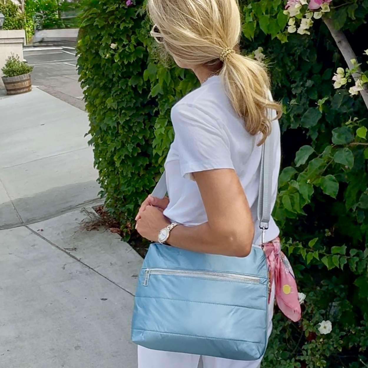 Woman standing near greenery carrying crossbody puffer purse in shimmer light blue