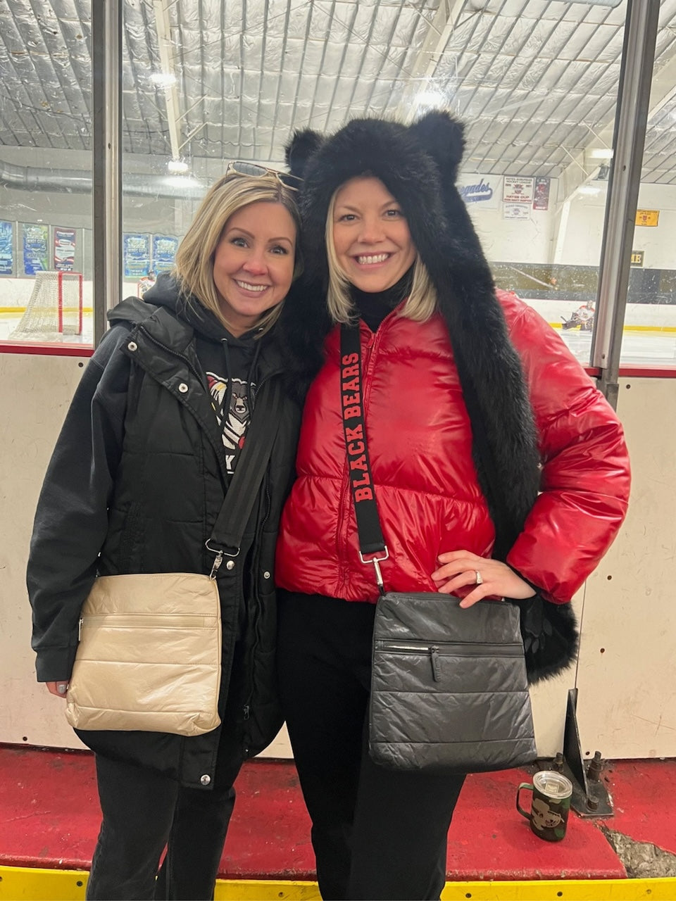 Two women standing together in an indoor ice rink area, one wearing a beige puffer purse and another a black crossbody purse