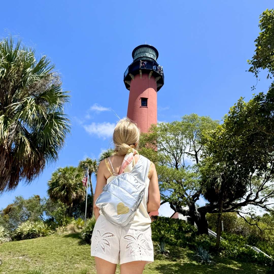 A woman with a silver backpack with a gold heart standing in front of a lighthouse with palm trees and blue sky.