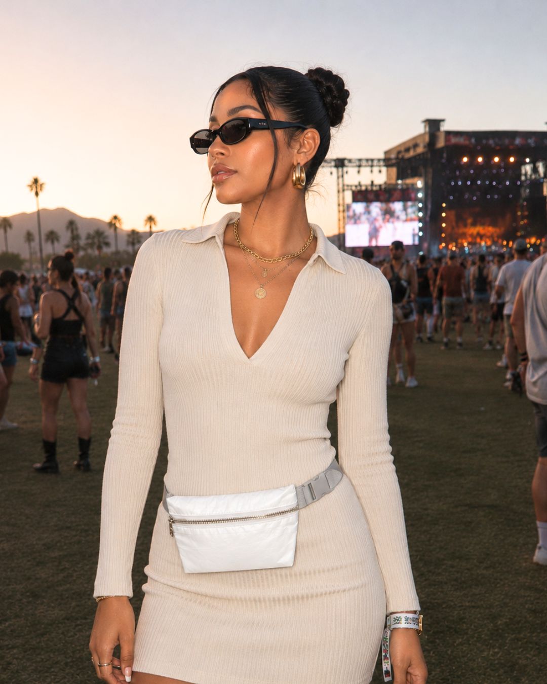 A woman at an outdoor festival wearing a cream ribbed long-sleeve mini dress with a collar, accessorized with a shimmer white belt bag, sunglasses, and gold jewelry.