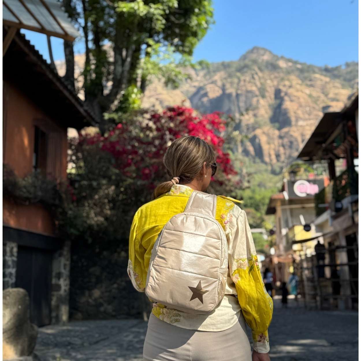 Person wearing a yellow shirt and shimmer beige backpack with a star design, standing in a scenic location with mountains and buildings.