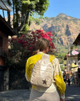 Person wearing a yellow shirt and shimmer beige backpack with a star design, standing in a scenic location with mountains and buildings.