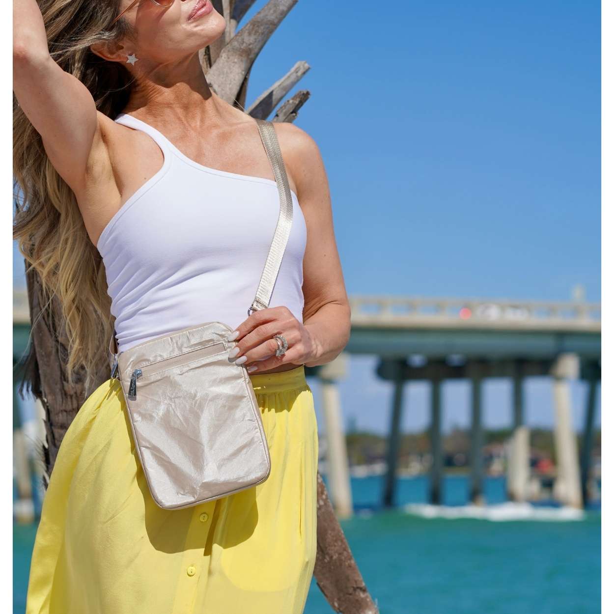 Woman in white top and yellow skirt with a beige handbag by a waterfront with a bridge in the background.