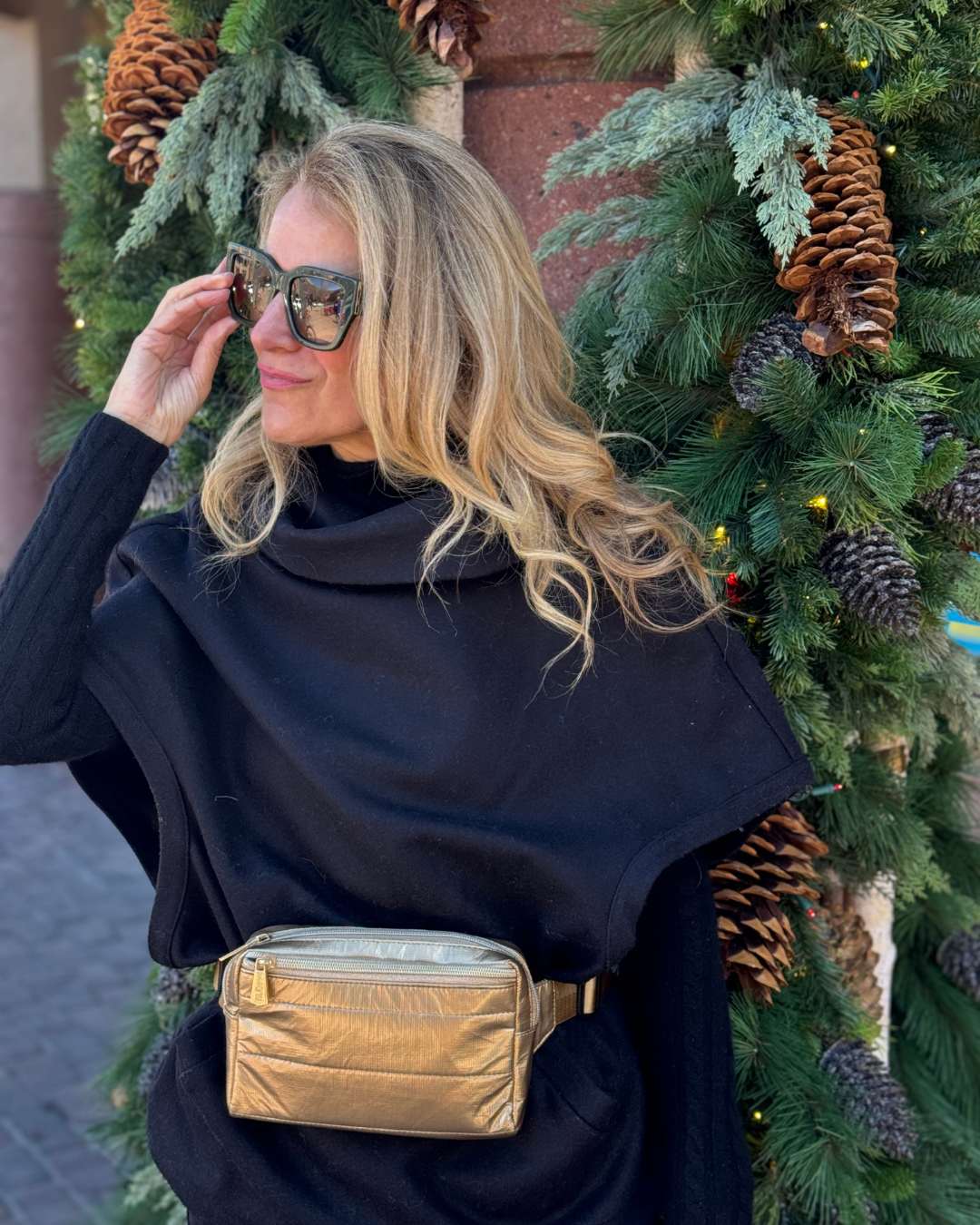 A woman wearing a black outfit with a gold crossbody fanny pack belt bag near holiday greenery