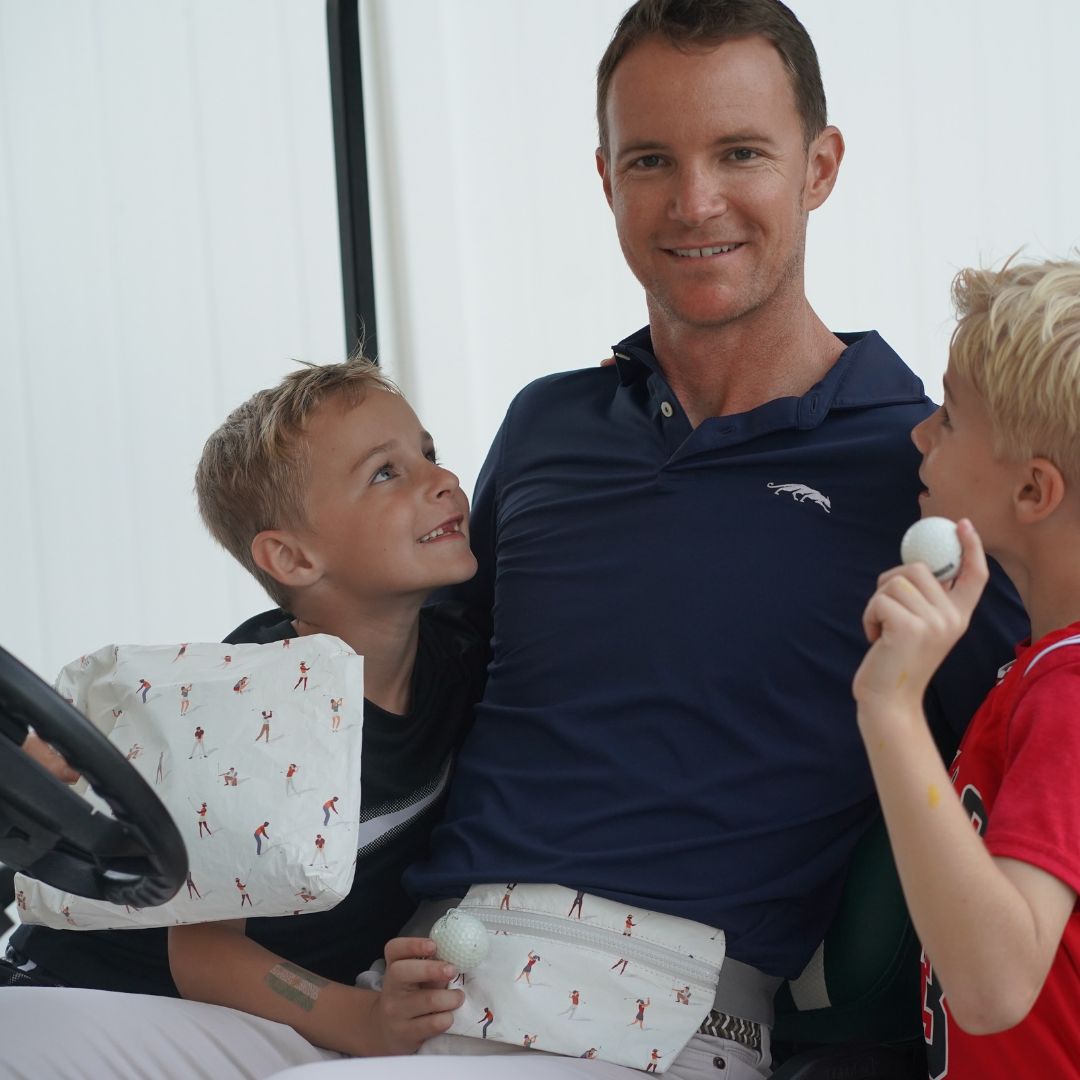 A man in a navy blue polo shirt sits in a golf cart with two young boys, holding golf balls wearing a golfer print belt bag and holding a golfer print zipper pouch. The background is a clean white wall.