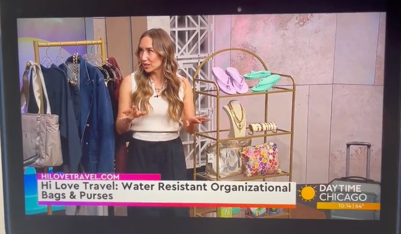 A television screen showing a Daytime Chicago segment with a woman speaking about water-resistant organizational bags and purses. The set includes clothing racks, jewelry stands, and travel bags, with on-screen text reading 'Hi Love Travel: Water Resistant Organizational Bags & purses' and a 'Daytime Chicago' logo.