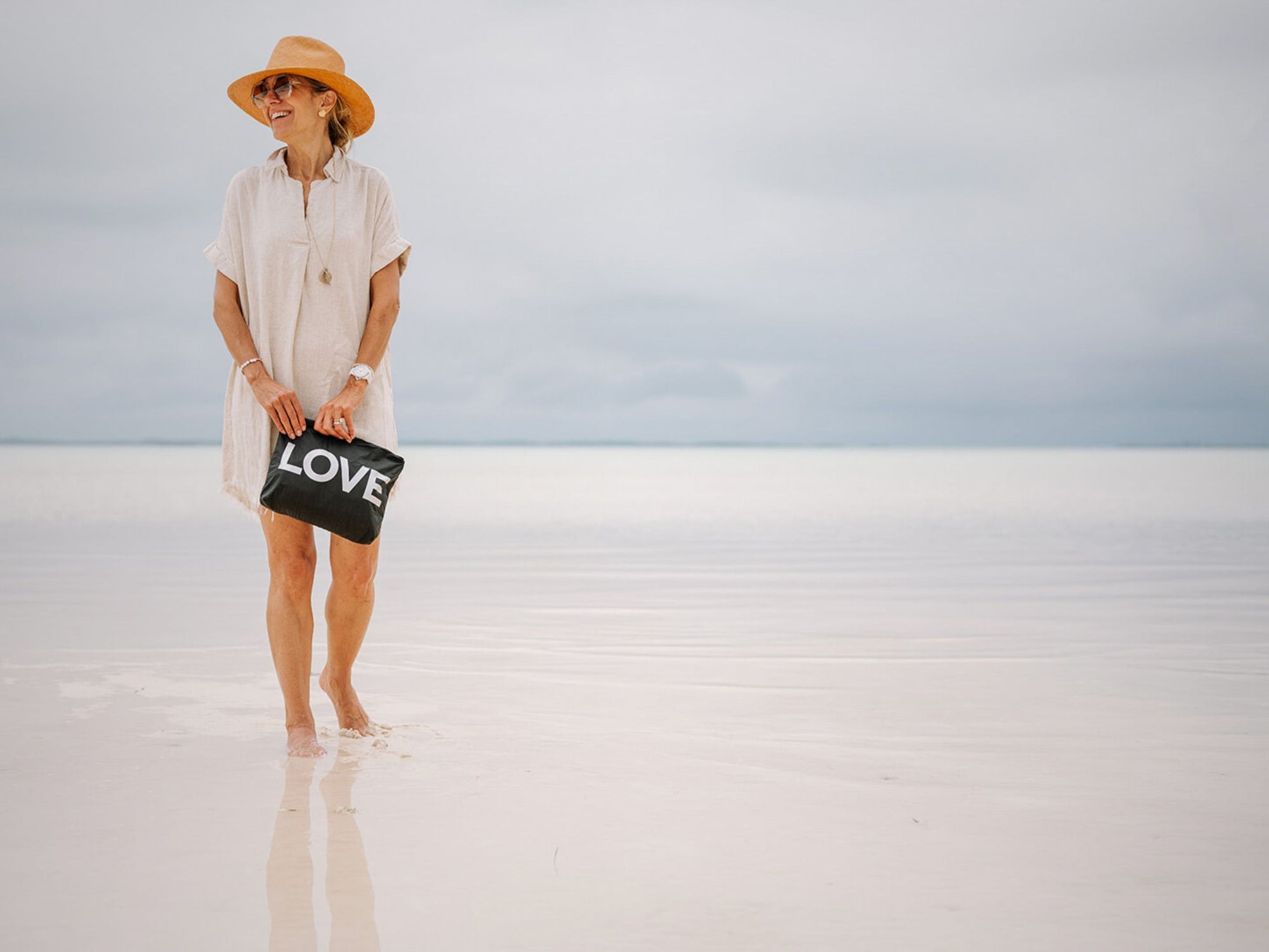 Woman holding a 'LOVE' zipper pouch on a reflective water surface with a clear sky.