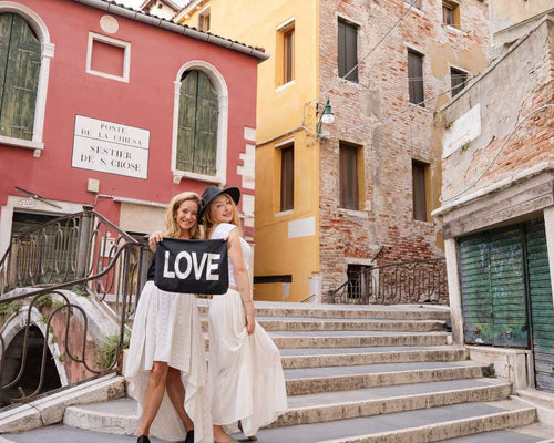 Two women holding a 'LOVE' zipper pouch in an Italian street with colorful buildings.