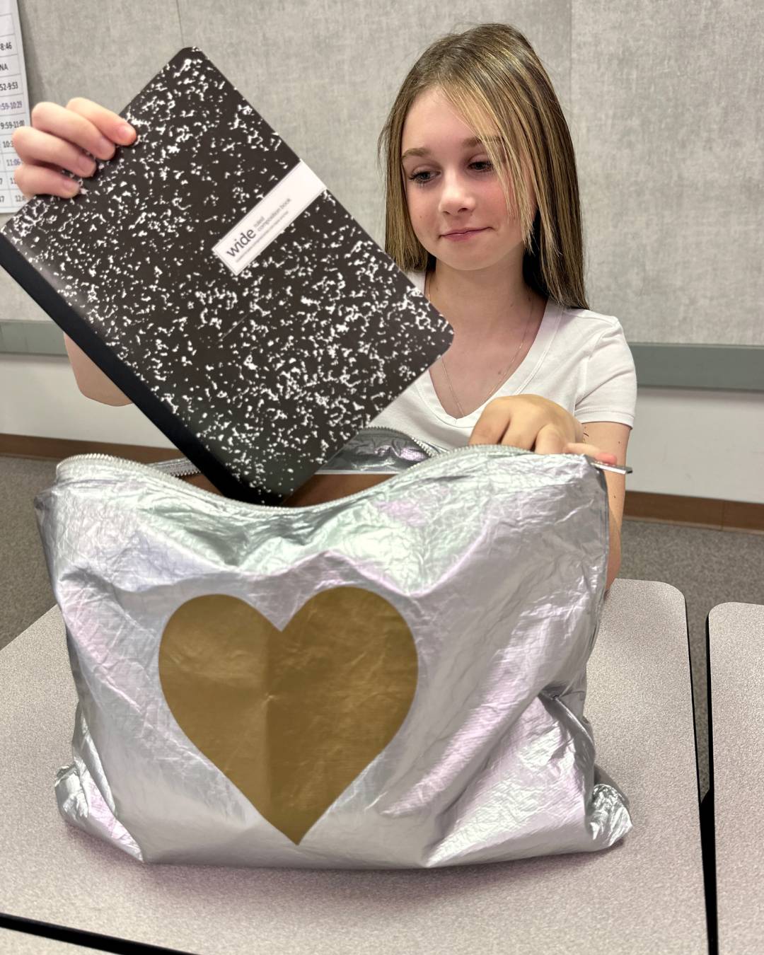A young girl with blonde hair sits at a desk holding a black-and-white speckled composition notebook, putting it into a large silver zipper pouch with a gold heart on the front.