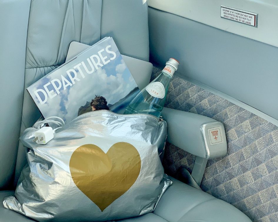 Silver bag with a gold heart, bottle, and 'Departures' magazine on an airplane seat.