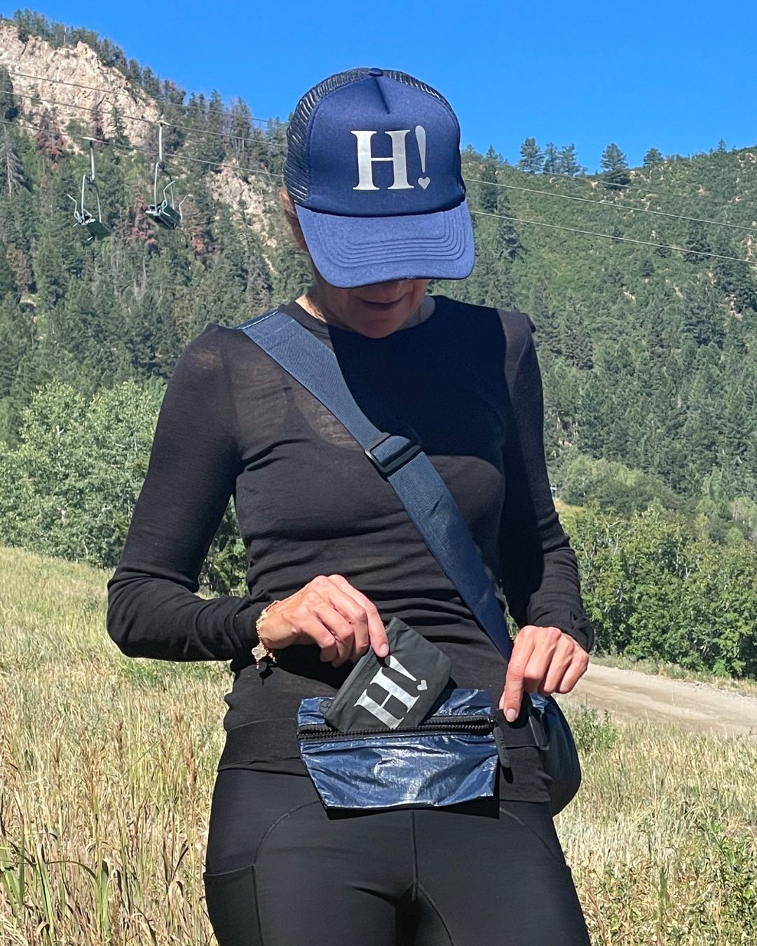 Person in field wearing black outfit with navy blue hat, lay flat fanny pack in navy blue, and card pouch in black with silver H!