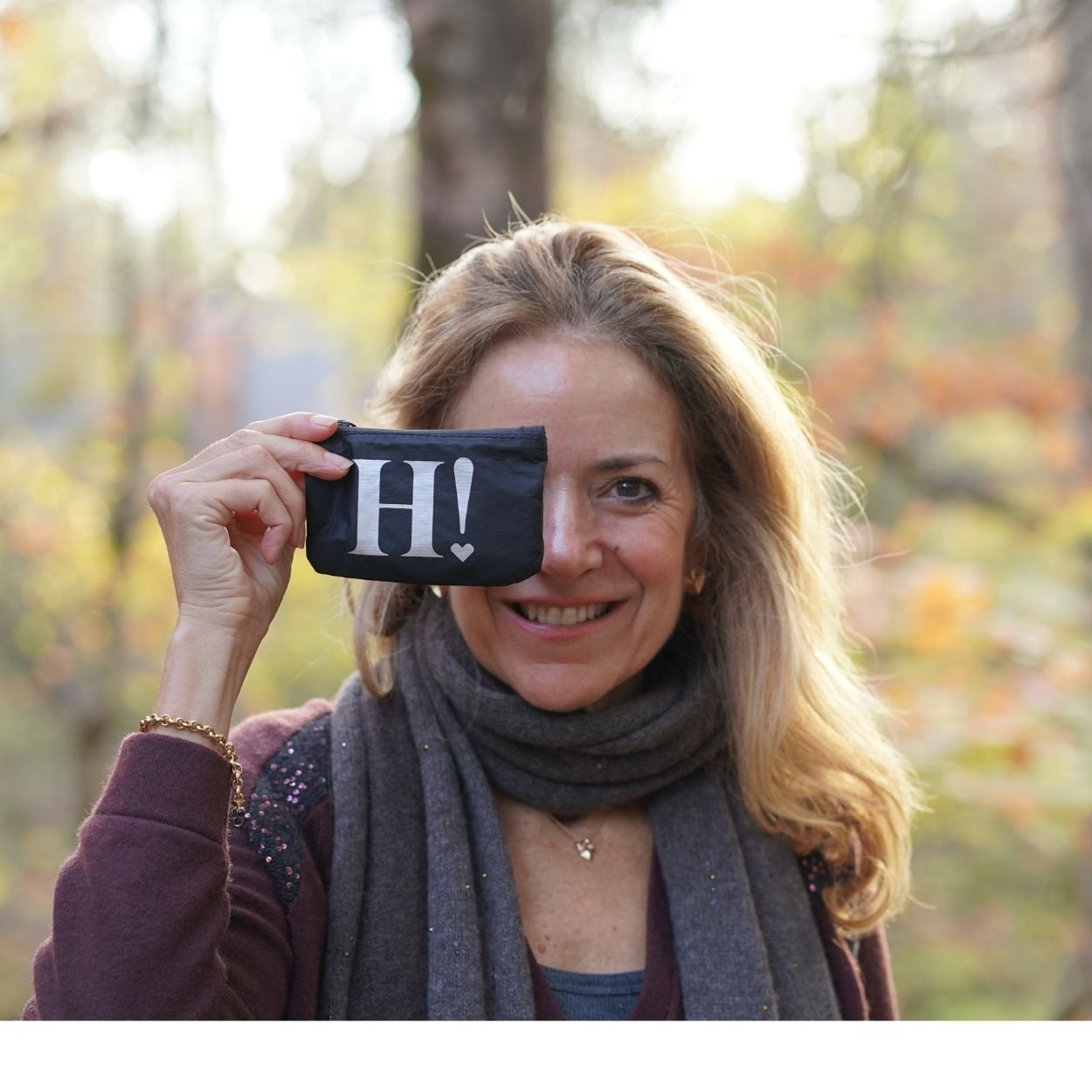 A smiling woman outdoors holds a small black pouch or case in front of her eye, which has the white letter 'H' and a small heart printed on it. She is wearing a gray scarf and a burgundy top, with a blurred autumn forest background.
