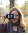 A smiling woman outdoors holds a small black pouch or case in front of her eye, which has the white letter 'H' and a small heart printed on it. She is wearing a gray scarf and a burgundy top, with a blurred autumn forest background.