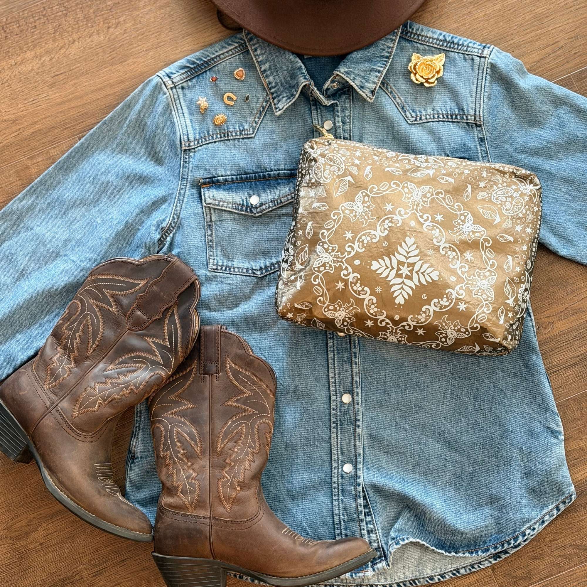 Medium Shimmer Bronze Bandana Print zipper pouch styled on a denim shirt beside brown cowboy boots and brown hat.