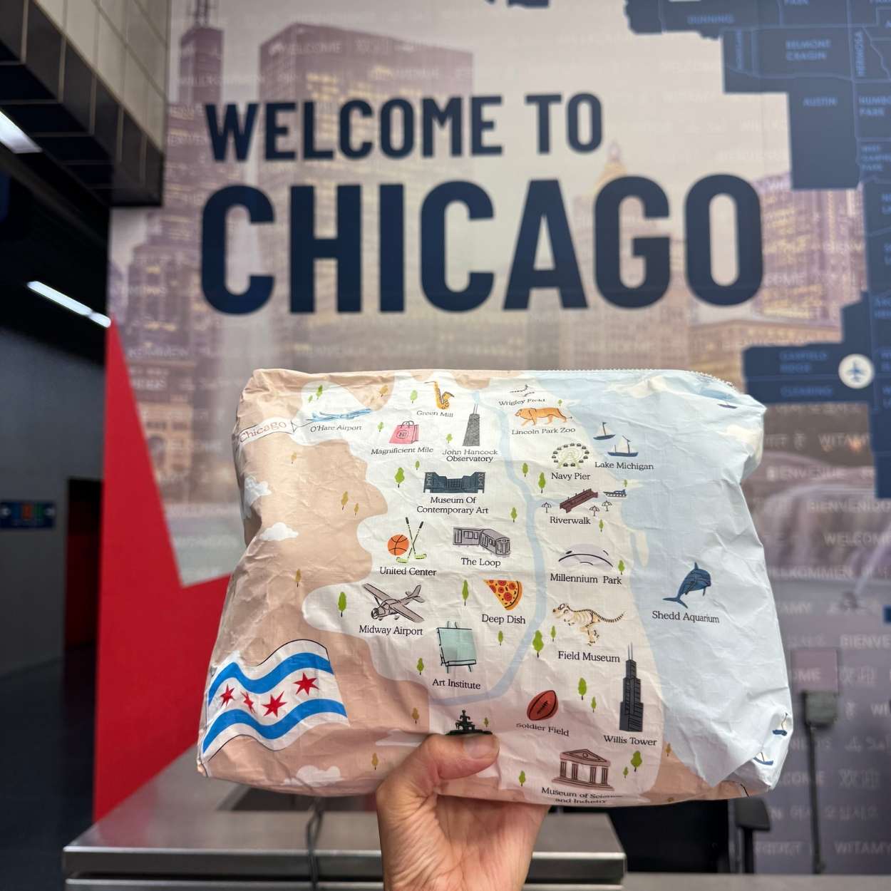 A hand holds a flat, rectangular printed pouch featuring a Chicago-themed illustrated map and city icons, with a Chicago flag in the lower left, in front of a large 'WELCOME TO CHICAGO' wall sign.