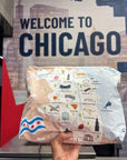 A hand holds a flat, rectangular printed pouch featuring a Chicago-themed illustrated map and city icons, with a Chicago flag in the lower left, in front of a large 'WELCOME TO CHICAGO' wall sign.