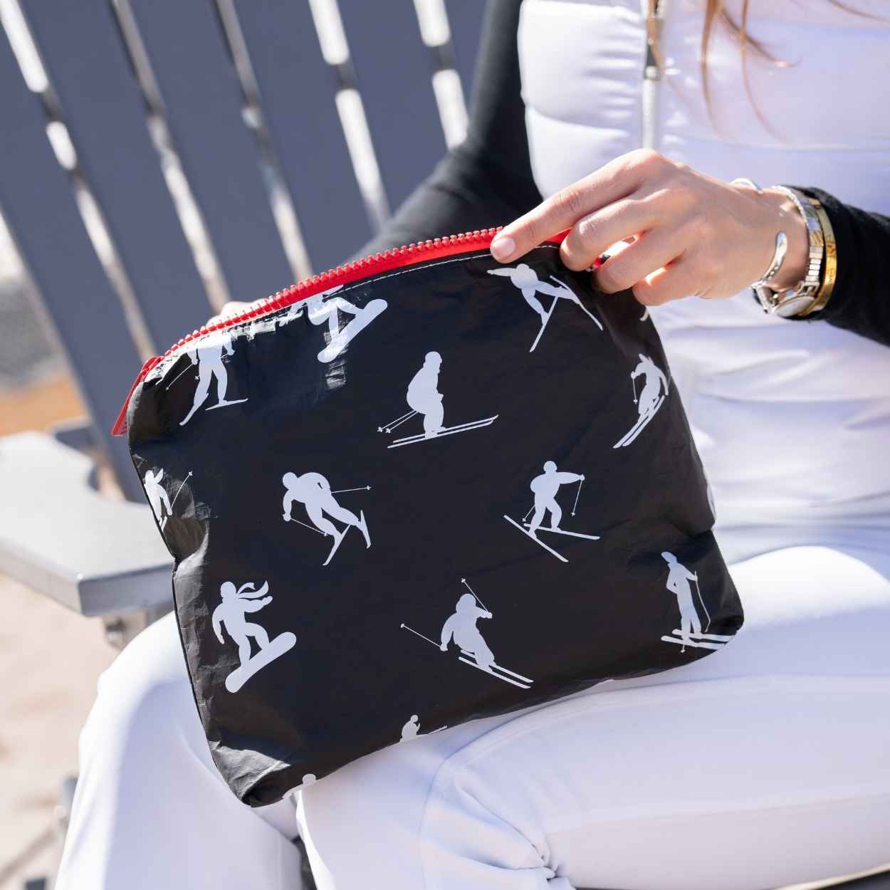 A black zippered pouch with a white ski and snowboard silhouette pattern and a red zipper, held by a person wearing white ski pants and a white top in an outdoor setting.