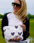 A woman outdoors holding a white zip pouch decorated with a pattern of gray and black watercolor hearts. She is wearing a white t-shirt, a black cardigan, and black sunglasses, with a green field in the background.