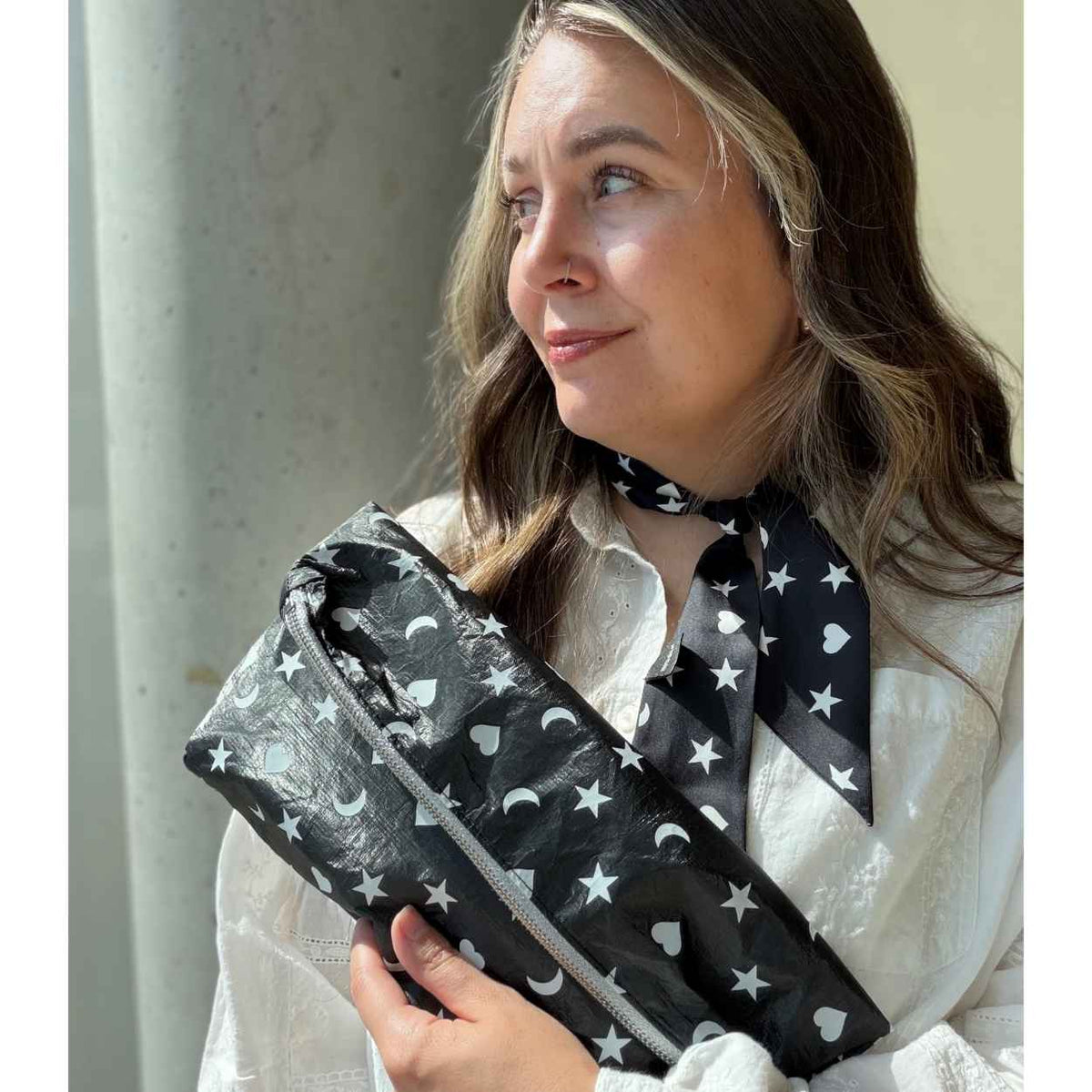 Woman in white top holding black clutch bag with white hearts, moons, and stars and wearing matching neck scarf against gray background