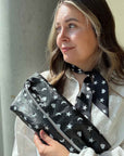 Woman in white top holding black clutch bag with white hearts, moons, and stars and wearing matching neck scarf against gray background