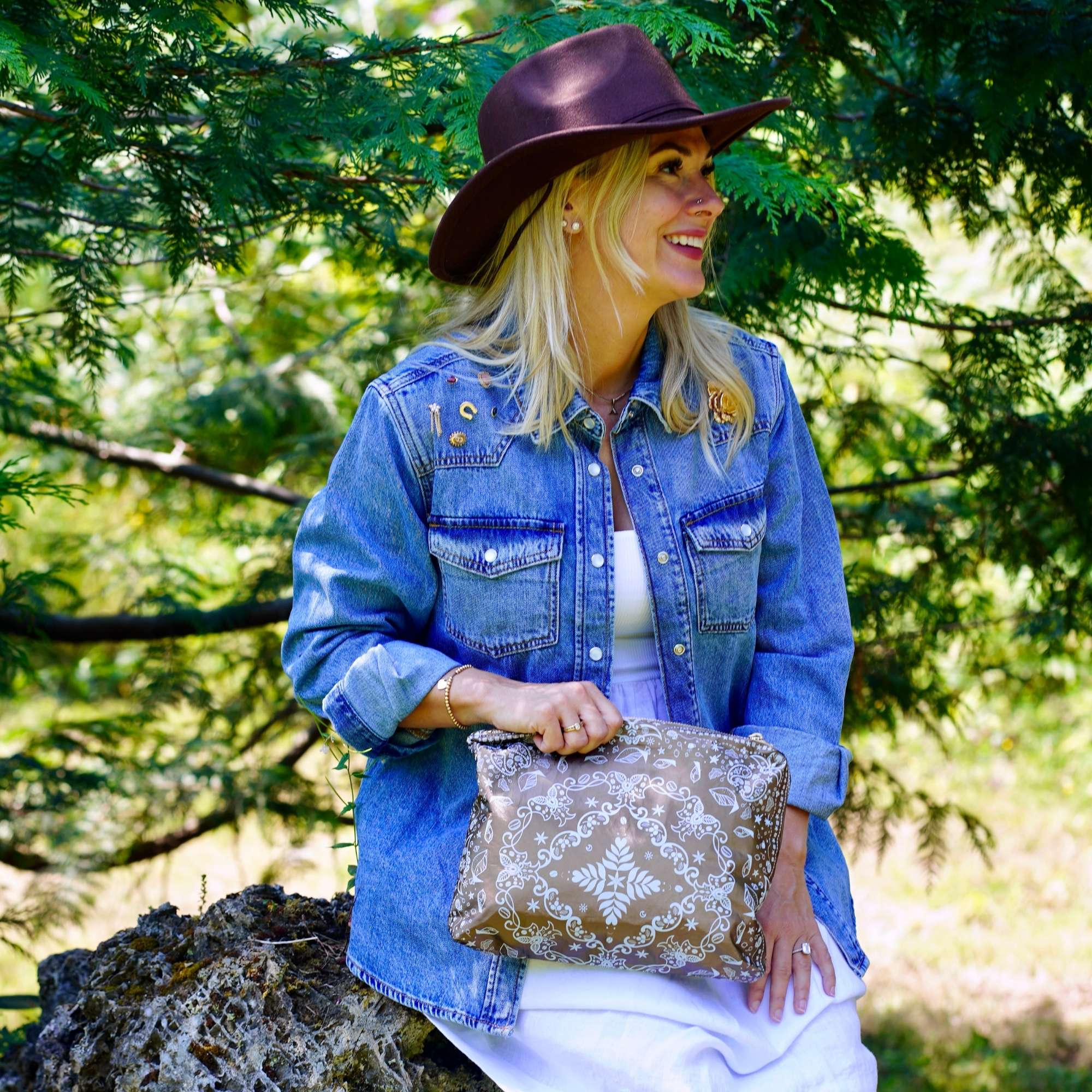 Woman in a denim shirt and hat sitting outdoors with a Medium Shimmer Bronze Bandana Print zipper pouch.