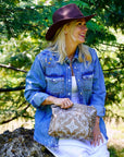 Woman in a denim shirt and hat sitting outdoors with a Medium Shimmer Bronze Bandana Print zipper pouch.