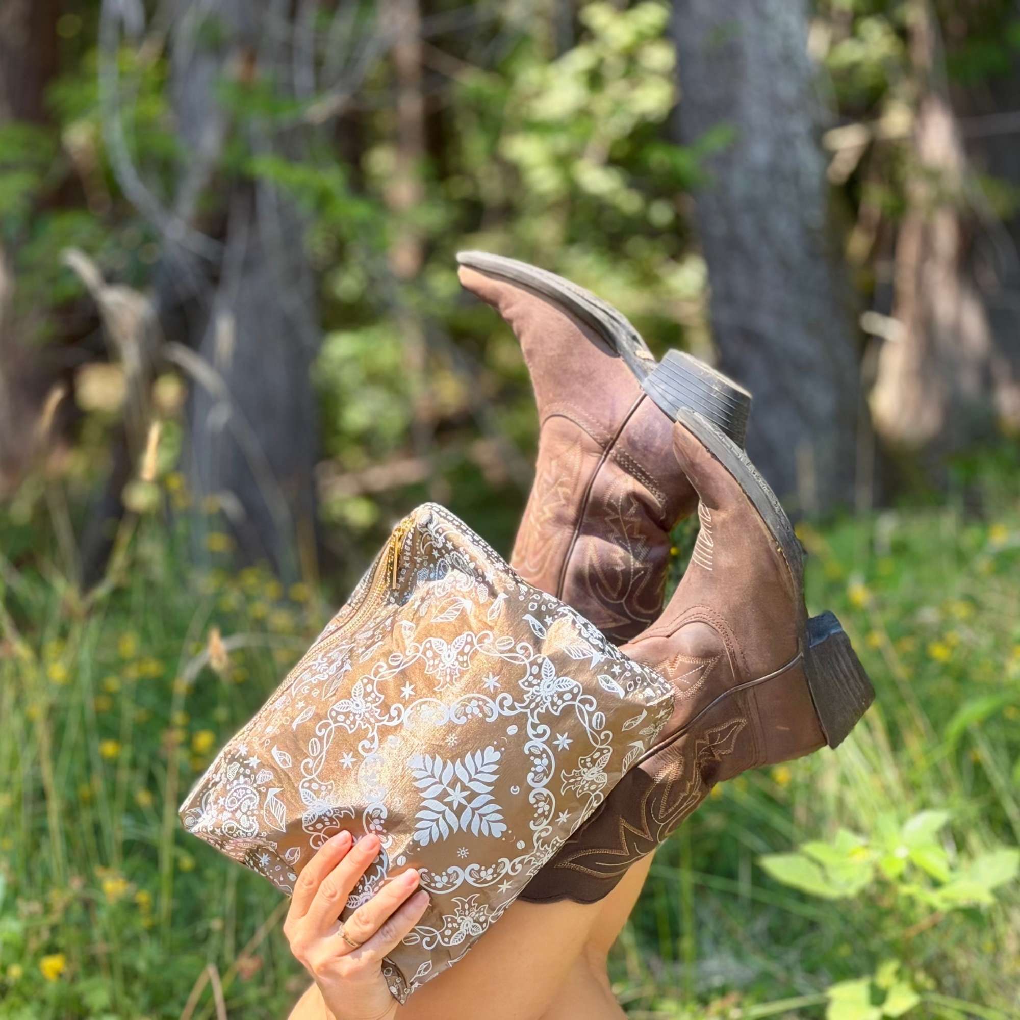 Medium Shimmer Bronze Bandana Print zipper pouch held beside brown cowboy boots in an outdoor setting.