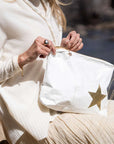 A person outdoors holding a white pouch with a large gold star and a small gold star charm, wearing a cream ribbed sweater and pleated cream skirt.