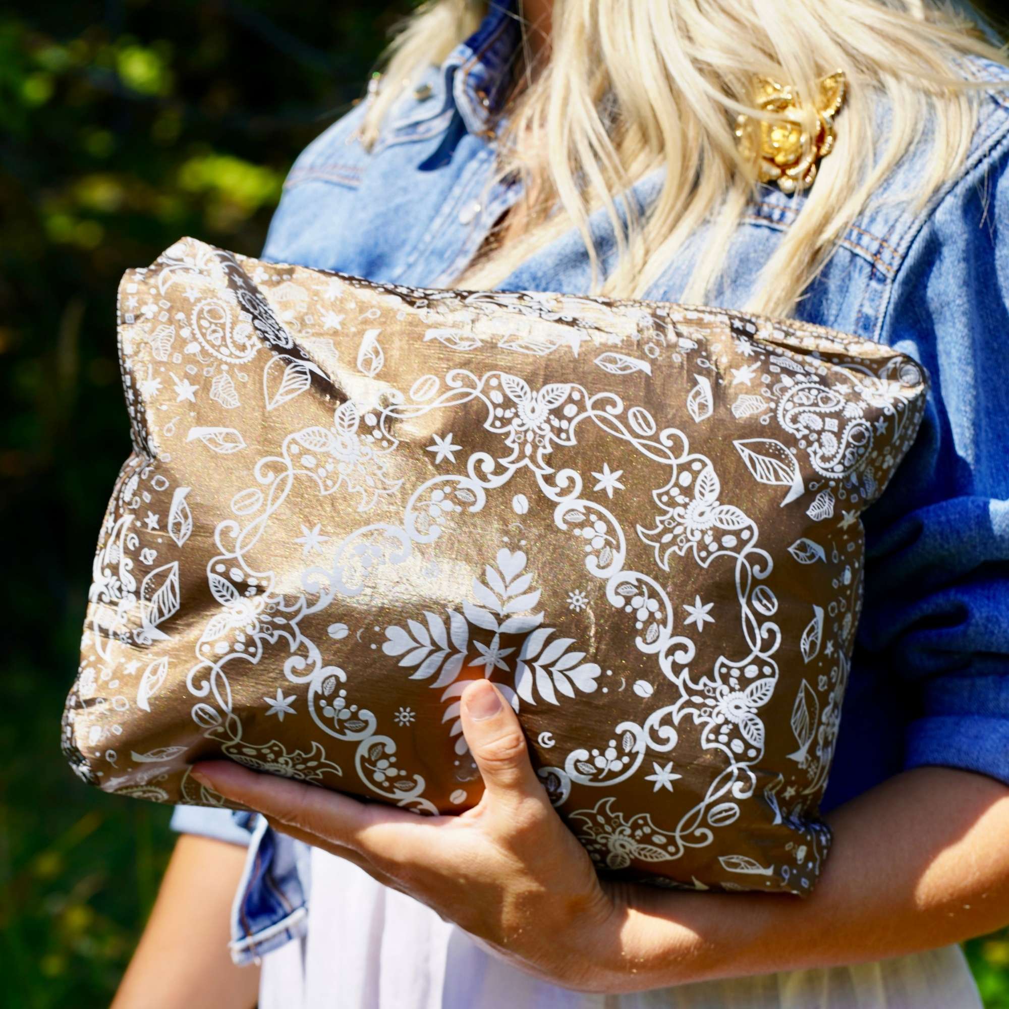 Woman in a denim shirt holding a Medium Shimmer Bronze Bandana Print zipper pouch outdoors.