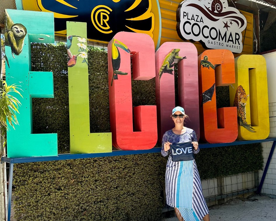 Woman in swimsuit and towel holding black pouch with love on it in Coco Beach Costa Rica in front of colorful signage