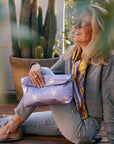 Woman sitting outdoors holding a purple clutch with star pattern, surrounded by plants.