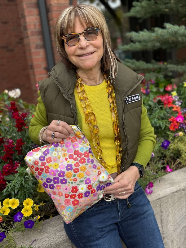 Woman holding a colorful, floral-patterned zipper pouch outdoors with flowers and a building in the background
